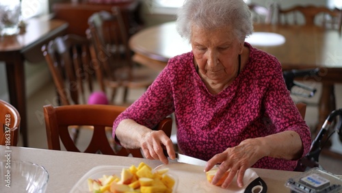 Slika na platnu Elderly senior woman slices fruit to make a salad while talking on a phone video