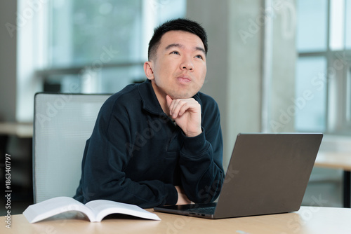 An Asian student looks upward thoughtfully with hand under chin while sitting at a desk with an open textbook and laptop, ideal for study break, brainstorming, or creative thinking concepts.