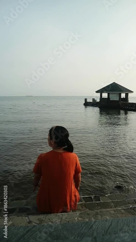 Woman in orange sitting on steps by the water looking out at a pier with a gazebo at dusk.