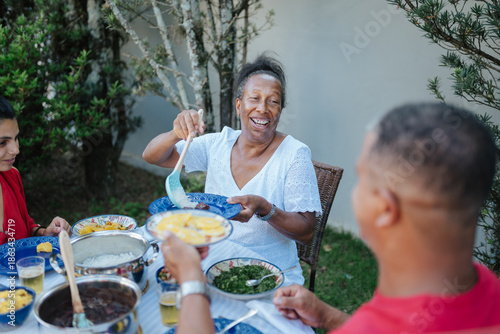 Konstfotografi Happy Senior Black Woman Serving Traditional Brazilian Feijoada to Family During