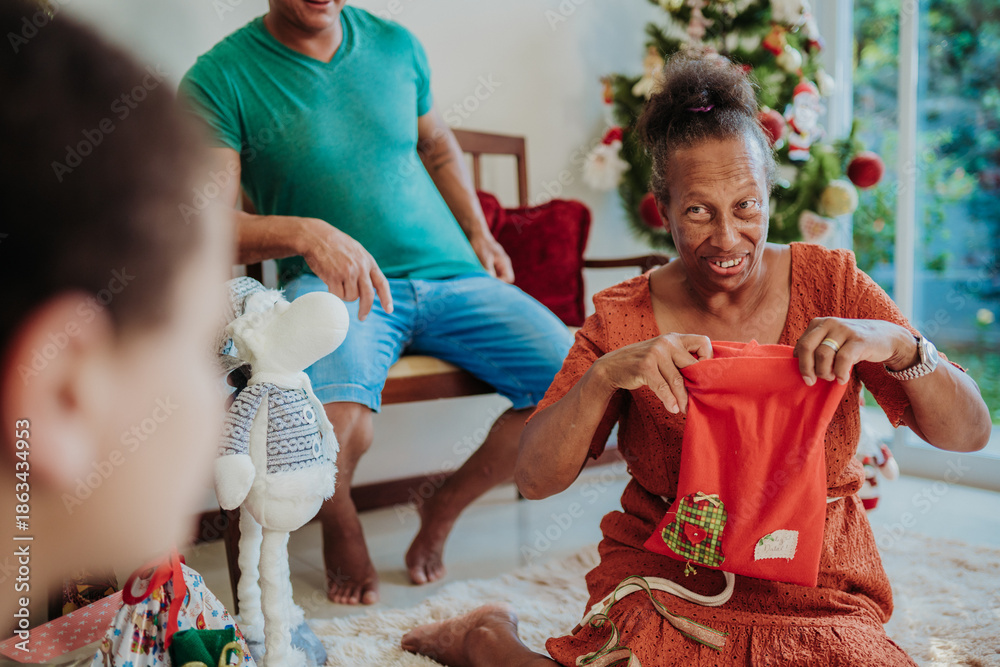 Obraz premium Happy senior Black woman opening a red Christmas gift bag while celebrating with her family at home near a decorated tree.