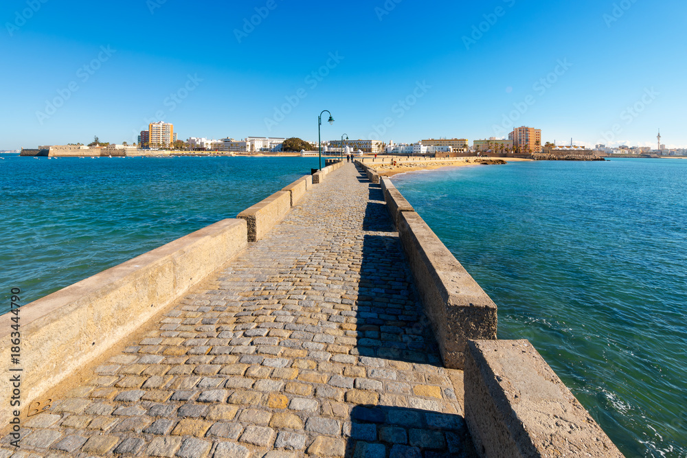 Fototapeta premium View of the sandy beaches and seaside city from the historic stone bridge walkway, Paseo Fernando Quiñones, that connects La Caleta beach to the Castle of San Sebastian, in Cadiz, Spain.