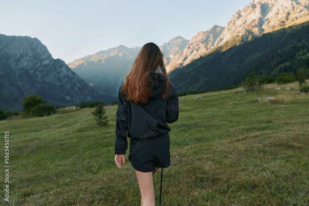 Naklejka premium A lone hiker walks through a lush alpine meadow, seen from behind. Dressed in a dark jacket and shorts, she explores vast mountains, exuding serenity and curiosity in a spacious outdoor setting.