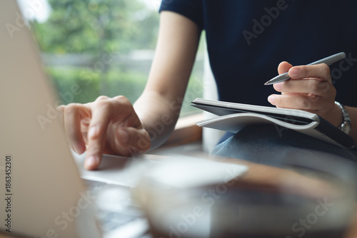Business woman sitting at table in cafe, writing in notebook working on laptop computer on table, doing online research. 