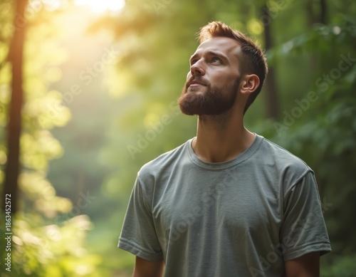 Man with beard looks up in sunlit forest. He feels calm and peaceful enjoying nature and fresh air. Mindful moment outdoors, person breathes deeply.