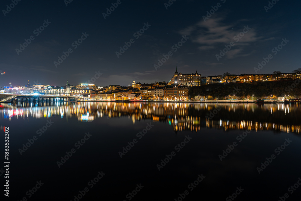 Obraz premium Nighttime skyline of Stockholm, Sweden, reflecting across calm water with illuminated historic buildings and city lights creating a glowing waterfront cityscape.