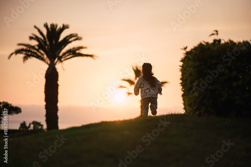 
A girl is running outdoors among bushes and palm trees, with the beach on the horizon. 