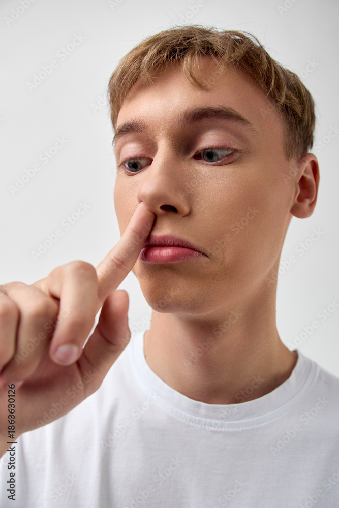 Fotografie Closeup of a casual young man in a white tshirt pressing his finger to a nostril in a humorous, candid moment, minimal indoor light, simple background, and a focused, self aware expression