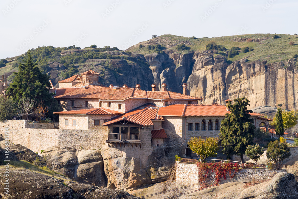 Obraz premium Traditional stone monastery complex with terracotta rooftops sits atop rugged sandstone rocks in Meteora, Greece, surrounded by greenery and dramatic cliffs in warm daylight.