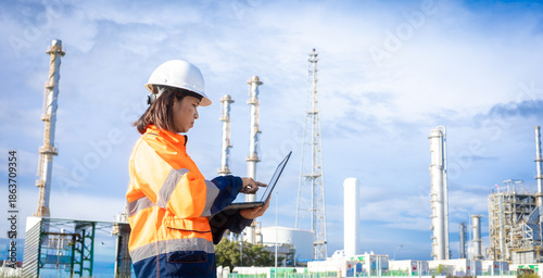 A worker stands outdoors at an industrial site. The person uses a laptop while wearing a safety helmet and bright clothing. Tall structures are visible in the background