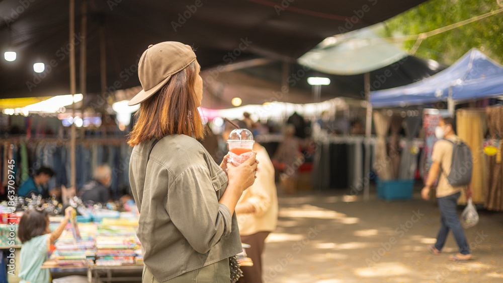 Fototapeta premium A woman stands with a drink in her hand looking at vendors in a busy market. Many people walk around shopping. Tents and stalls surround the area. It is a sunny day