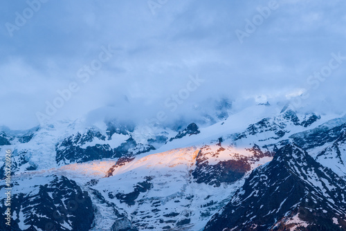Valokuva Warm sunlight briefly illuminates a section of snow-covered Mont Blanc du Tacul, contrasting against dramatic blue shadows and swirling clouds in the high alpine landscape