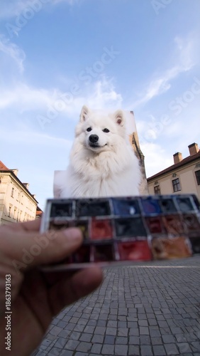 A person holds watercolors while a fluffy white dog portrait towers above on a sunny cobblestone plaza