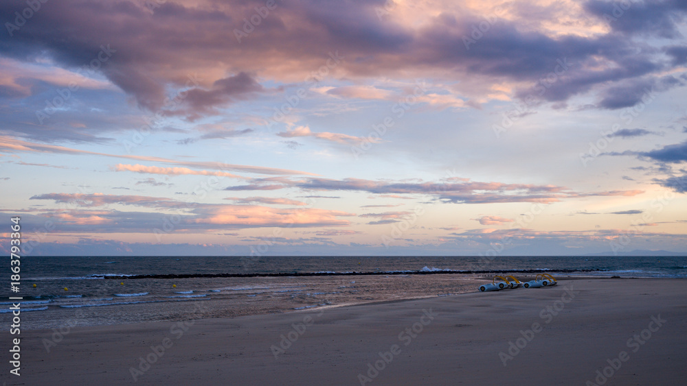 Fototapeta premium Soft pastel clouds stretch across the evening sky above a deserted sandy beach in Agde, with gentle waves and a row of yellow pedal boats adding subtle detail to the tranquil coastal scene.