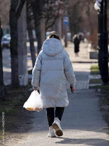A woman with a package is walking along the sidewalk in the rays of sunlight. Pedestrian on a city street. Everyday life of a woman mistress at home. Shopping trip to the store. Using a plastic bag