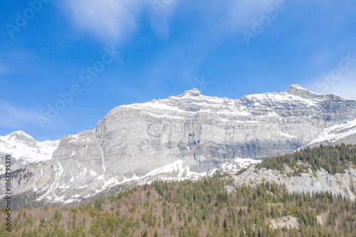Slika na platnu Expansive view of the dramatic Pointe de Plate limestone cliffs rising above a dense alpine forest, with patches of lingering snow and a vivid blue sky overhead