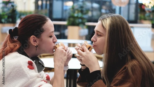 Two pretty girls eat burgers with gusto at a table in a fast food cafe.