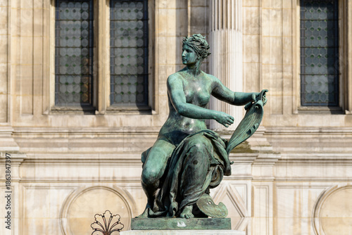 Photos Patinated bronze sculpture of a seated woman holding a scroll, set against the historic stone facade and tall windows of Hotel de Ville in Paris, illuminated by soft natural light
