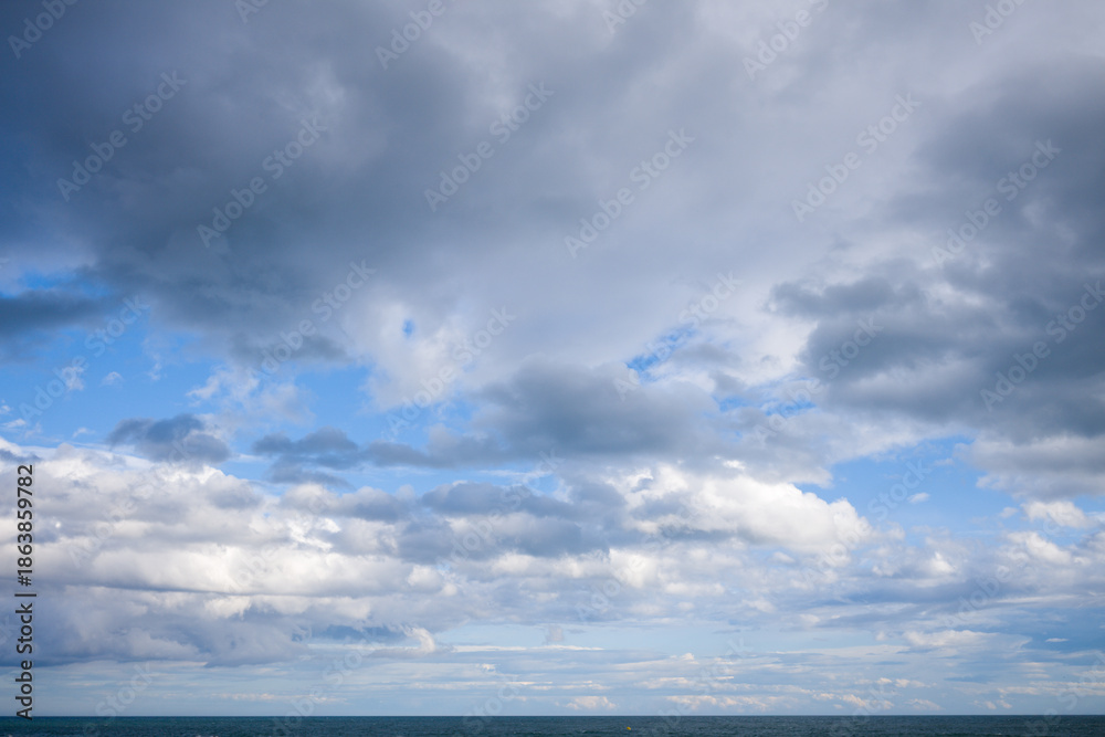 Fototapeta premium Vast cloud-filled sky stretches above a tranquil blue sea, with soft light illuminating the gentle waves near Agde. The scene evokes openness and serenity under shifting weather.