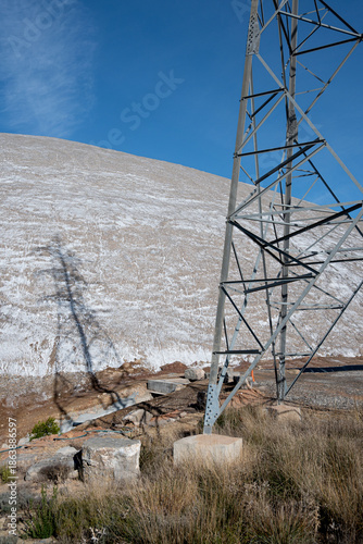 A towering metal lattice electricity transmission pylon stands prominently on the right, its robust structure framed against a bright blue sky with subtle wispy clouds. 
