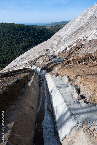 A steep hillside covered in white mineral deposits, likely salt or mining waste, dominates the foreground. A concrete drainage channel, or flume, with a thin stream of liquid runs down the slope