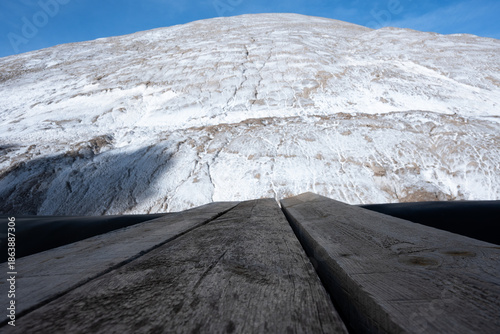 Massive white salt mountain under blue sky, viewed from weathered wooden planks. Symbolizes raw natural resources, grand challenges, aspiration, and a unique perspective on the journey ahead.