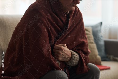 Senior Caucasian man sitting on sofa wrapping himself in blanket, while being sick with cold or flu, or unable to afford home heating, showing visible wrinkles and wearing glasses