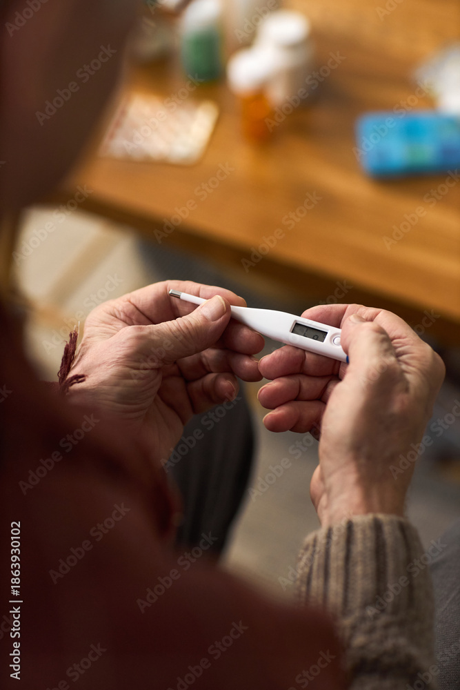Obraz premium Senior Caucasian man holding digital thermometer with both hands, checking temperature while sitting at table with medication bottles and blister packs in background