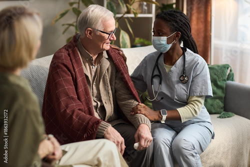 Tablou pe pânză Senior Caucasian man sitting on sofa being comforted by Black female nurse weari