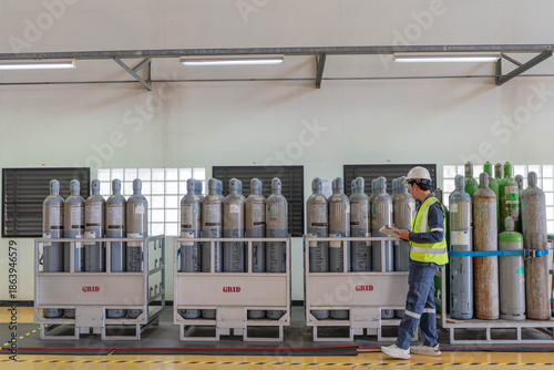 Professional male engineer in safety uniform walking and inspecting rows of industrial gas cylinders in warehouse, conducting workplace safety audit and inventory management in factory.