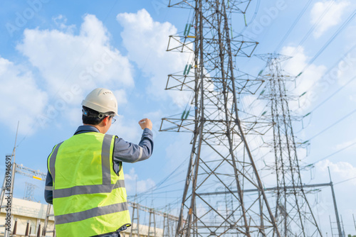 Back view of electrical engineer raising hand to point at high voltage transmission towers at power substation, supervising grid infrastructure development and energy supply maintenance.