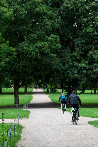 Two people cycling leisurely along a vigeland park path through lush green trees on a sunny summer day. Oslo, Norway