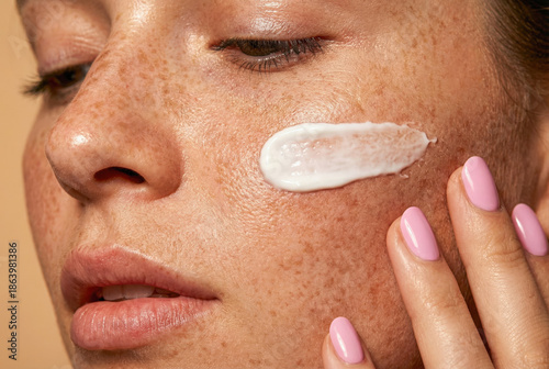 Close up of young woman with natural freckled skin applying white moisturizing cream on her cheek, skincare and beauty concept
