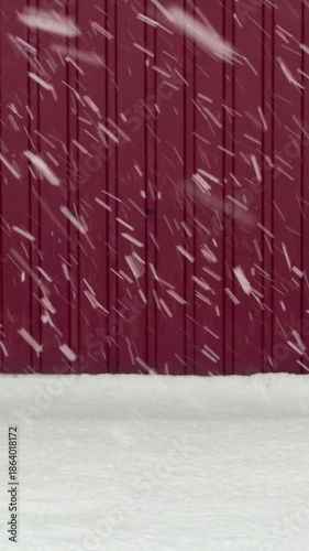 The snow is falling in a beautiful, swirling pattern against the backdrop of a red wall. The snowflakes are large and white, creating a stunning contrast against the warm, rich color of the wall