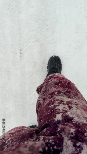 A person is walking in the snow wearing a red coat and black boots. The boots are covered in snow, and the person's feet are visible. The snow is falling lightly, and the person is walking on a pat
