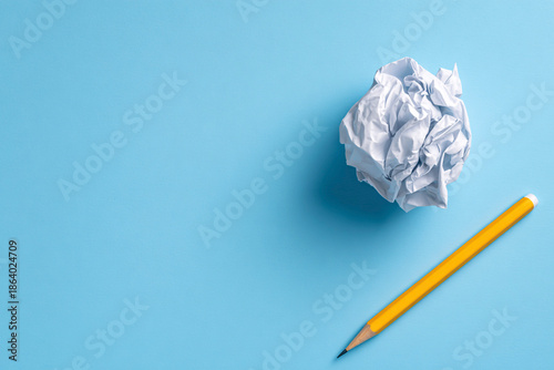 Top view flat lay of crumpled white paper ball and yellow pencil on blue background representing writer's block and creativity