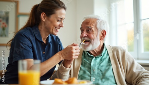 Caregiver feeds happy elderly man breakfast at home or care facility. Smiling nurse helps senior with food, provides support and nourishment. Healthy senior enjoys meal assisted by woman.