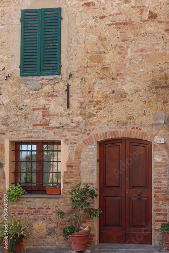 A charming entrance in Montepulciano, Tuscany, featuring a rustic stone wall, a wooden door, and a window with green shutters. Beautiful plants add a touch of warmth to this Italian countryside scene.