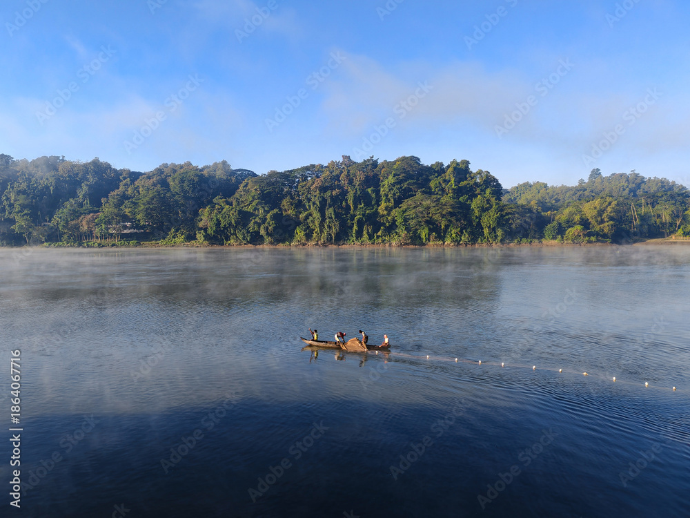 Fototapeta premium A wide shot of a tranquil river under clear blue sky captures a small wooden fishing boat carrying people navigating the calm waters, and a fishing wall of tropical forest lines the far bank