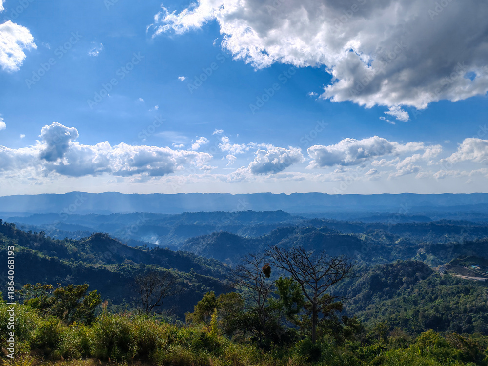 Obraz premium Panoramic view overlooking rolling green hills and distant blue mountain ranges under bright blue sky with dramatic white clouds covering horizon