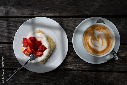 A strawberry biscuit and a cup of cappuccino are on a wooden table.
