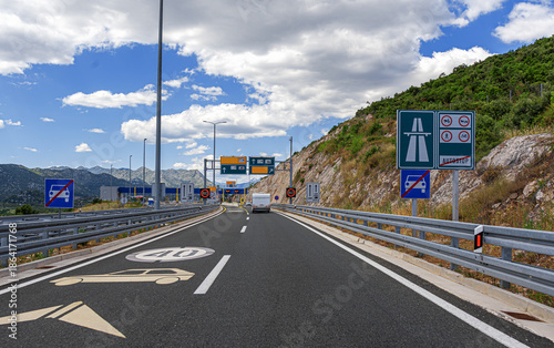 Fotografie A car with a trailer approaches a highway toll station and border crossing nestled in a mountainous landscape under a blue sky with white clouds