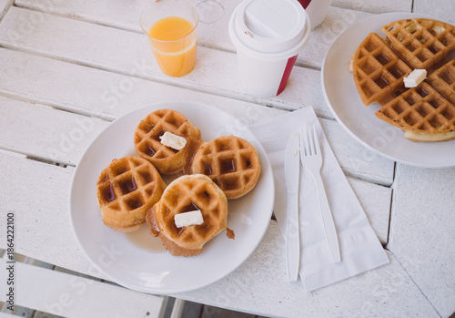 Typical American hotel or motel breakfast: waffles with butter and maple syrup, orange juice and coffee in disposable takeaway cups, plastic cutlery on white outdoor patio table