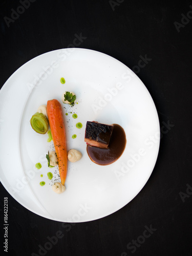 Slow cooked pork belly with celeriac puree, glazed carrot and leek served in fine dining gourmet restaurant. White porcelain plate on black background. Art of plating food.