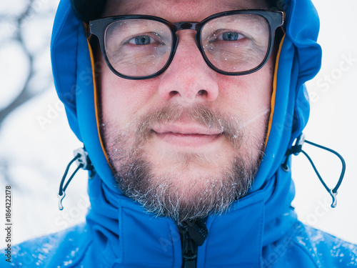 Close up of smiling face of middle-aged man wearing glasses and blue outdoor jacket with hoodie during snow storm with snowflakes in his beard, male with frames outside during cold winter