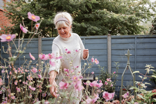 Elderly woman in a white sweater and floral skirt picking flowers in a summer garden, calm and relaxed atmosphere with soft natural light.