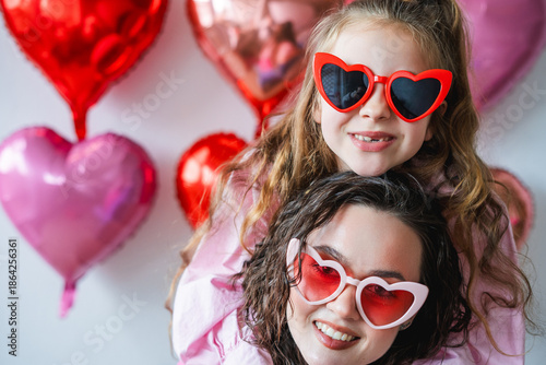 Cute little girl with her teenage sister in heart glasses on a background of pink balloons. Valentine's Day.