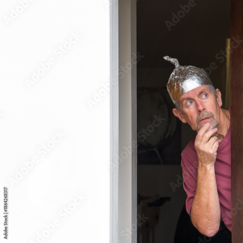 Tinfoil hat picture. A man wearing a tinfoil hat looks anxiously out of a door towards the sky. Paranoia and conspiracy theory. Alien abduction and government cover up. Mind control and brainwashing. 