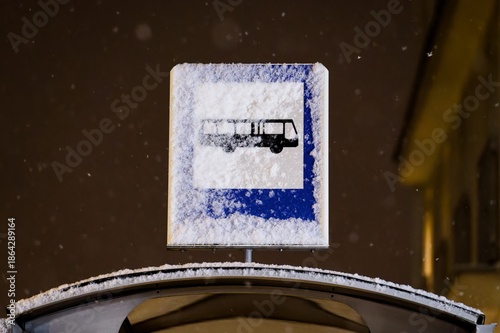 Snow covered bus stop sign at night with falling snow, winter public transportation scene and cold weather conditions.