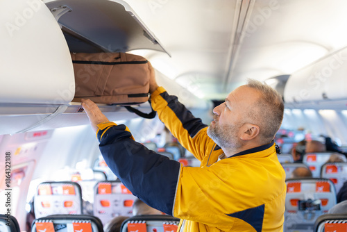 Quadro em tela Adult caucasian man placing carry on backpack into overhead bin inside airplane cabin